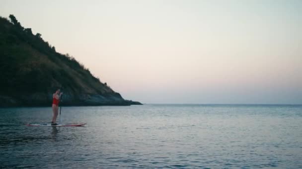 Mujer en traje de baño rojo en SUP de pie tabla de paddle en una bahía al atardecer — Vídeo de stock