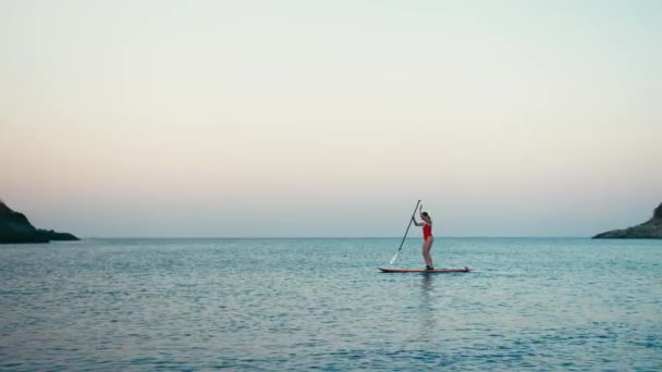 Женщина в красном купальнике на SUP встала на весло доску на заливе на закате — стоковое видео