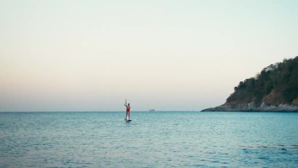 Femme en maillot de bain rouge sur SUP debout planche à pagaie sur une baie au coucher du soleil — Video