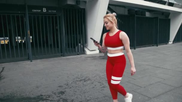 Más tamaño rubia mixta racee sonriente mujer usando ropa deportiva roja usando teléfono inteligente al aire libre — Vídeo de stock