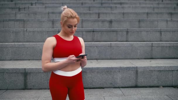 Além de tamanho loiro misto de corrida sorrindo mulher vestindo sportswear vermelho usando smartphone ao ar livre — Vídeo de Stock