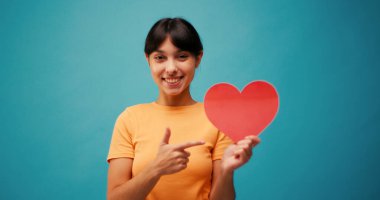 Neşeli İspanyol kadın mavi arka planda parlak bir gülümsemeyle kalp sembolü gösteriyor. Kadın, sevgiyi anlamlı jestlerle gösterir. Yavaş çekim