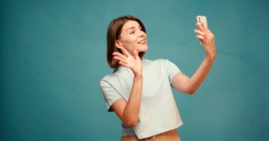 İspanyol kadın el sallıyor ve mavi arka plandaki akıllı telefonu kullanarak görüntülü konuşuyor. Kadın dalgaları görüntülü konuşma sırasında mutlu oluyor. Yavaş çekim