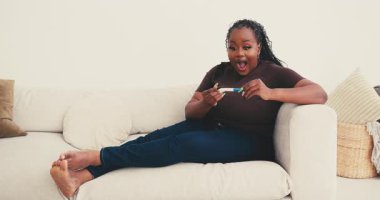 Afro-Amerikalı bir kadın hamile testi için koltukta oturuyor ve gülümsüyor. Olumlu sonuçtan sonra neşeli tepkiler ortaya çıkıyor. Kadın zıplarken çığlık atıyor