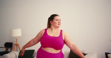 Şişman Artı Beden Kadın yoga minderinde savaşçı pozuyla kollarını açıyor. Kilo verme sürecinde, parlak oturma odasında spor sutyen egzersizi yapan kadın.