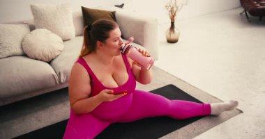 Spor kıyafetleri giyen kilolu artı beden kadın elinde su şişesi olan telefon çekleri tutuyor. Odada antreman yaptıktan sonra su molası verilir.