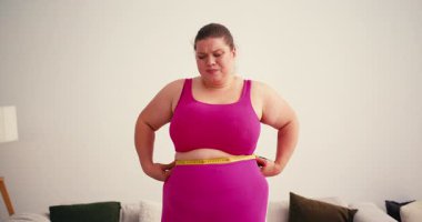 Kıvrımlı, kilolu ve spor giyimli kadın, bel çevresini ölçmek için mezura kullanıyor. Kadın kilo vermeyi hayal eden ölçülere bakıyor.