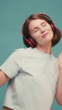 Şık kulaklıklı İspanyol kadın müziğin ritmini seviyor ve mavi arka planda zarif bir şekilde dans ediyor. Mutlu kadın neşeli melodiyi sever. Yavaş çekim