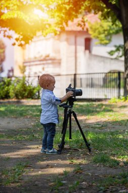 İki yıl yaşlı erkek çocuk fotoğrafçıdır. Çocuk bir fotoğraf makinesi tripod üzerinde tutan ve manzara fotoğrafı çeker. Küçük çocuk gömlek ve parkta oynarken kot giymiş.