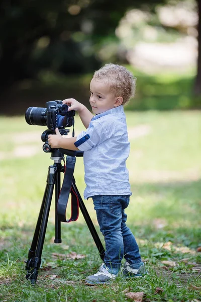 两岁的男孩是摄影师 孩子拿着三脚架上的相机 拍下风景 小孩子穿衬衫和牛仔裤在公园里玩耍 — 图库照片