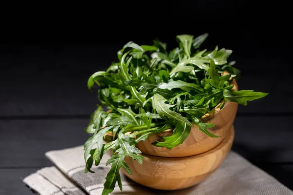 Fresh rucola in the wood bowl full of rocket salad leaves over the black background, selective focus