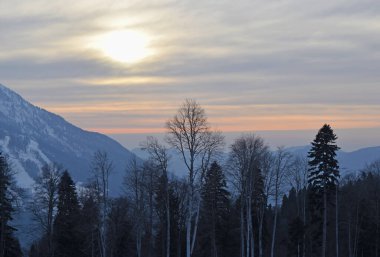 Karlı, yüksek dağlar ve ormanın üzerinde bulutlu bir gökyüzünde güzel bir kış günbatımı.