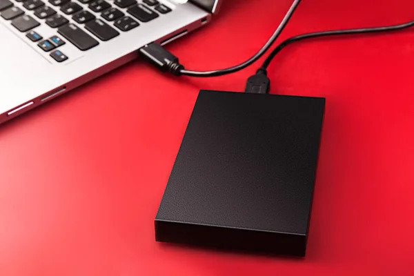 An external hard drive connected to the laptop with a usb cable on a red background. Portable storage technologies