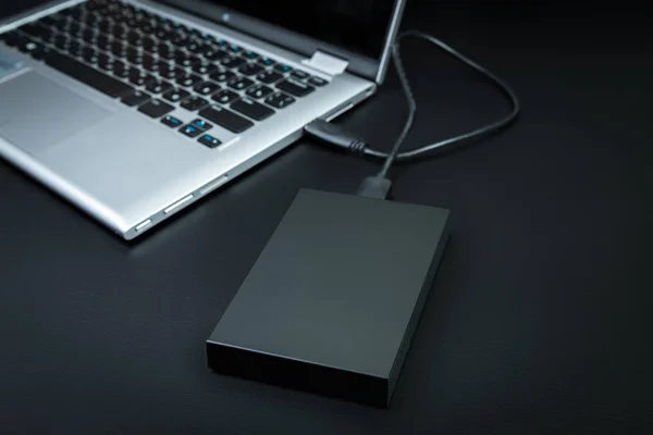 An external hard drive  connected to the laptop with a usb cable on a black background. Portable storage technologies