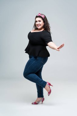 Siyah bluz ve mavi kotlu, gri stüdyo arka planda şişman bir kadın, vücut pozitifliği.