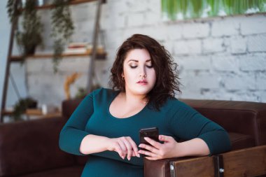 Artı yeşil elbiseli akıllı telefon kullanan bir model, evde şişman bir kadın, vücut pozitifliği.
