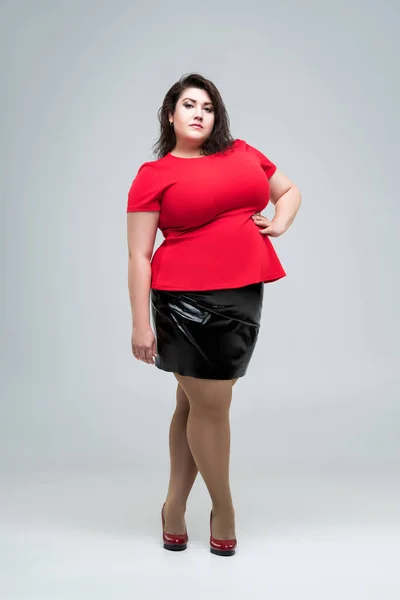 Plus size fashion model in red blouse and black skirt, fat woman on gray studio background, body positive concept