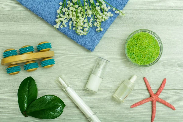 Syringe with eye cream, bottles with cream for face skin and aromatic oil, hand massager, bowl with sea salt, starfish and bouquet of lilies of the valley on blue towel. Spa products and accessories