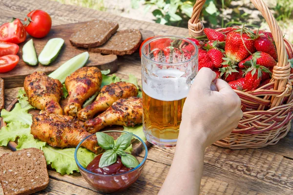 El cam bardak birayla ızgara tavuk budu ve domates, salatalık, siyah dehşet, hasır sepet çilek arka planda tutuyor. Kupa tarih seçici odak