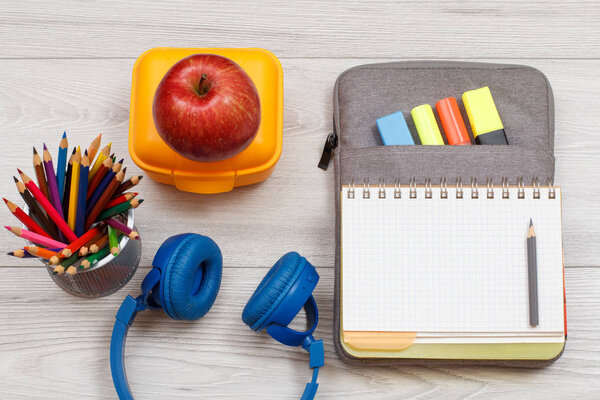 Headphones, metal stand with color pencils, yellow sandwich box, apple and open exercise book on bag-pencil case with color felt pens and marker on grey wooden boards. Top view. Back to school concept. School supplies.