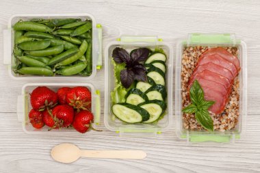 Plastik yemek hazırlık kapları ile taze çilek, yeşil bezelye, salatalık ve salata, haşlanmış Buğday lapası ve et, tahta kaşık gri arka plan üzerinde dilim. Üstten görünüm