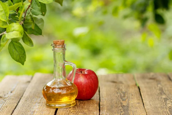 Apple vinegar in glass bottle with cork and fresh red apple on old wooden boards with blurred green natural background. Organic food for health