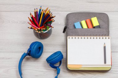 Kulaklıklar, metal renkli kalemler ile stand ve alıştırma kitabı çanta-kalem davasında hissettim renkli kalemler ve gri ahşap kurullarında marker ile açın. Üstten görünüm. Geri okul kavramı. Okul malzemeleri.