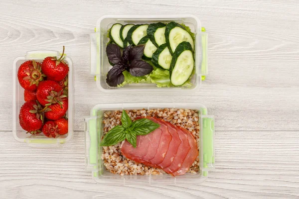 Plastik yemek kapları taze çilek, salatalık ve salata, haşlanmış Buğday lapası ve gri ahşap arka plan üzerinde et dilimleri ile hazırlayın. Kopya alanı ile üstten görünüm