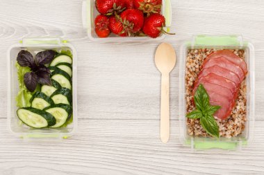 Plastik yemek kapları taze salatalık ve salata, haşlanmış Buğday lapası ve et, tahta kaşık gri ahşap zemin üzerine dilim çilek ile hazırlayın. Kopya alanı ile üstten görünüm
