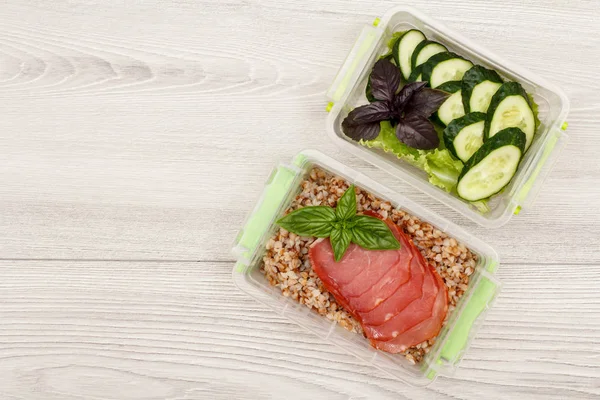 Plastic meal prep containers with boiled buckwheat porridge and slices of meat, fresh cucumbers and salad on grey wooden background. Top view with copy space