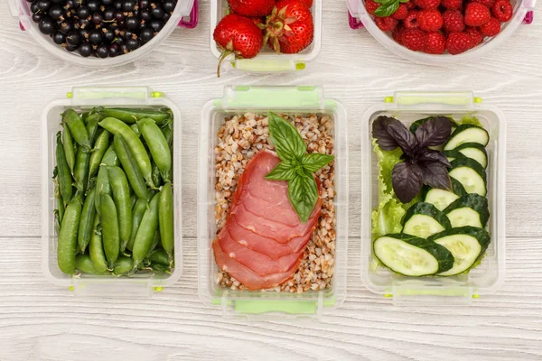 Plastic meal prep containers green peas, with boiled buckwheat porridge and slices of meat, fresh cucumbers and salad, currant, strawberry, raspberry on grey background. Top view