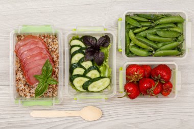 Plastik yemek kapları haşlanmış Buğday lapası ve dilim et, taze salatalık ve salata, çilek, yeşil bezelye ve gri arka plan üzerinde tahta kaşık ile hazırlayın. Üstten görünüm