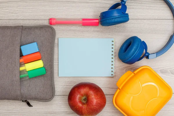 Çanta-kalem çalışmasıyla renk kalemler ve marker, elma, gri ahşap zemin üzerine mavi defter, kulaklık ve öğle yemeği kutusu hissettim. Üstten görünüm kopya alanı ile. Geri okul kavramı. Okul malzemeleri.