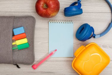 Çanta-kalem çalışmasıyla renk kalemler ve marker, elma, gri ahşap zemin üzerine mavi defter, kulaklık ve öğle yemeği kutusu hissettim. Üstten görünüm kopya alanı ile. Geri okul kavramı. Okul malzemeleri.