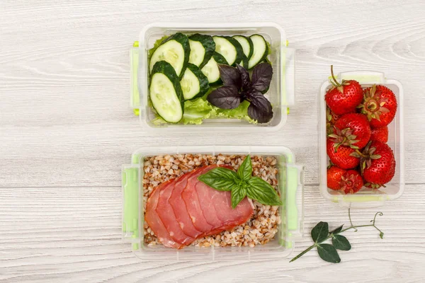 Plastic meal prep containers with boiled buckwheat porridge and slices of meat, fresh cucumbers and salad, strawberries on grey wooden background. Top view with copy space