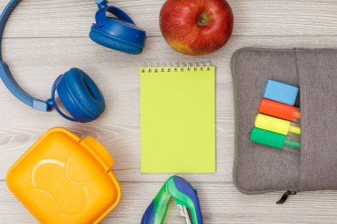 Beslenme çantası, kulaklık, zımba, sarı defter, elma ve çanta-kalem çalışmasıyla hissettim renkli kalemler ve marker gri ahşap zemin üzerinde. Üstten görünüm kopya alanı ile. Geri okul kavramı. Okul malzemeleri.