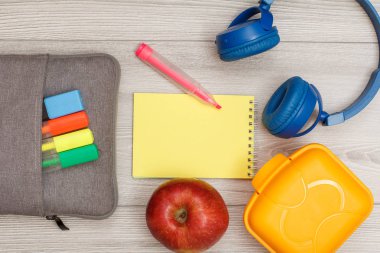 Çanta-kalem çalışmasıyla renk kalemler ve marker, elma, gri ahşap zemin üzerine sarı defter, kulaklık ve öğle yemeği kutusu hissettim. Üstten görünüm kopya alanı ile. Geri okul kavramı. Okul malzemeleri.