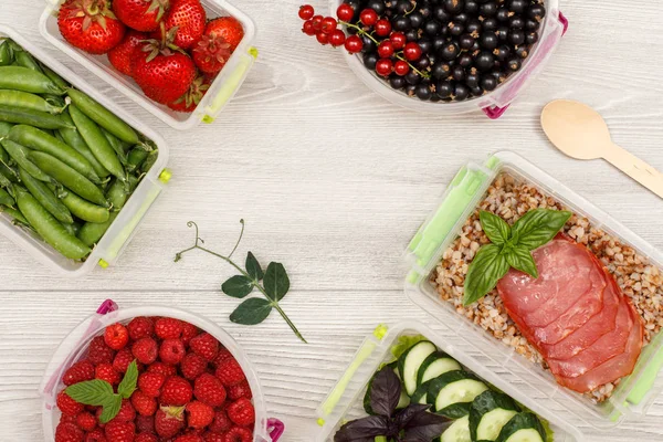 Plastic meal prep containers with green peas, strawberries, black currant, raspberries, fresh cucumbers and salad, boiled buckwheat porridge and slices of meat on grey background with copy space. Top view