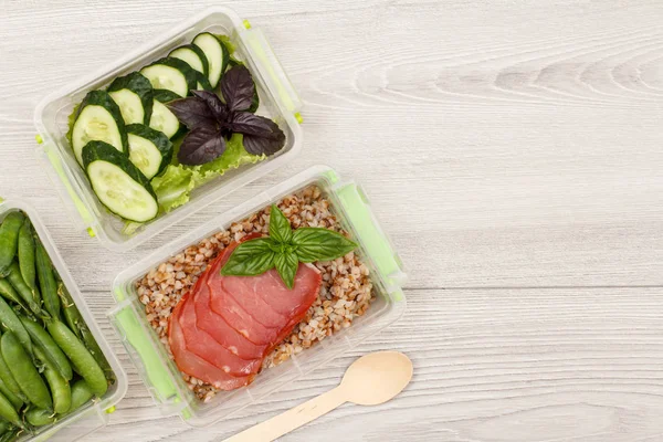 Plastic meal prep containers with green peas, boiled buckwheat porridge and slices of meat, fresh cucumbers and salad, wooden spoon on grey background. Top view with copy space