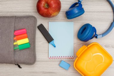 Çanta-kalem çalışmasıyla renk kalemler, elma, gri ahşap zemin üzerine mavi defter, kulaklık ve öğle yemeği kutusu hissettim. Üstten görünüm kopya alanı ile. Geri okul kavramı. Okul malzemeleri.