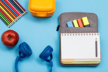 Apple, renkli kalemler, beslenme çantası, kulaklık, hissettim renkli kalemler ve mavi zemin üzerine marker ile egzersiz kitap çanta-kalem dava açın. Üstten görünüm. Geri okul kavramı. Okul malzemeleri.