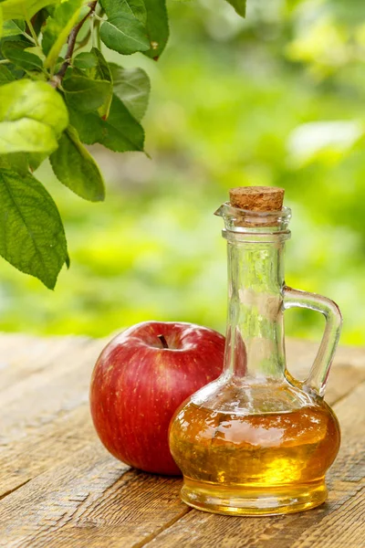 Apple vinegar in glass bottle with cork and fresh red apple on old wooden boards with blurred green natural background. Organic food for health