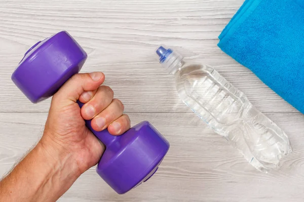 Male hand is holding a dumbbell in room or gym with bottle of water and towel in the background. Tools for fitness. Top view
