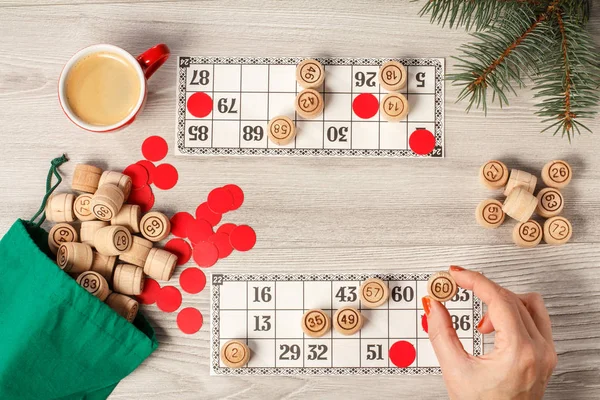 Woman's hand holding a barrel for a game in lotto. Wooden lotto barrels with green bag, game cards, red chips and cup of coffee, Christmas fir tree branches. Board game lotto. Top view