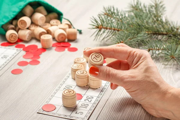 Woman\'s hand holding a barrel for a game in lotto. Wooden lotto barrels with green bag, game cards and red chips, Christmas fir tree branches on the background. Board game lotto