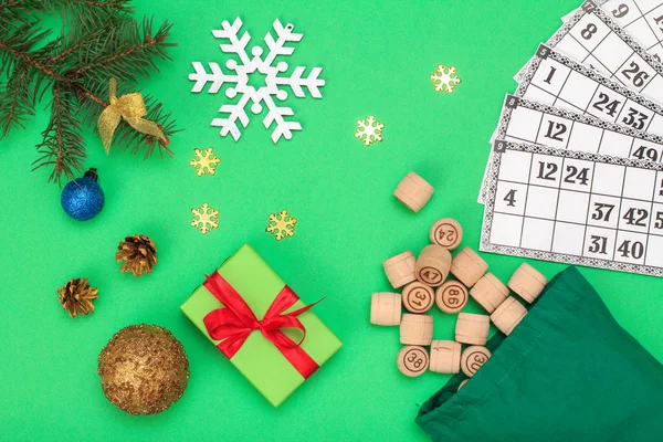 Board game lotto. Wooden lotto barrels with bag and game cards for a game in lotto, Christmas fir tree branches, cones, toy ball and gift box on green background. Top view