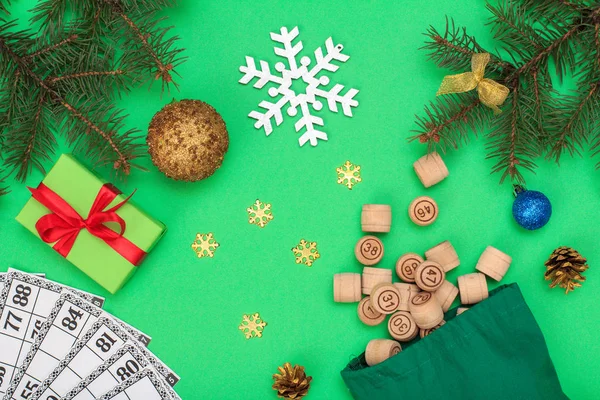 Board game lotto. Wooden lotto barrels with bag and game cards for a game in lotto, Christmas fir tree branches, cones, toy balls, snowflackes and gift box on green background. Top view