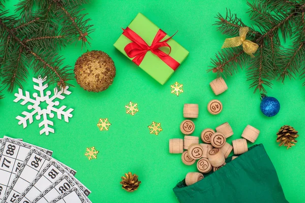 Board game lotto. Wooden lotto barrels with bag and game cards for a game in lotto, Christmas fir tree branches, cones, toy balls, snowflackes and gift box on green background. Top view