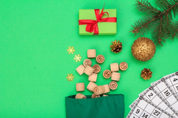 Board game lotto. Wooden lotto barrels with bag and game cards for a game in lotto, Christmas fir tree branch, cones, toy ball and gift box on green background. Top view