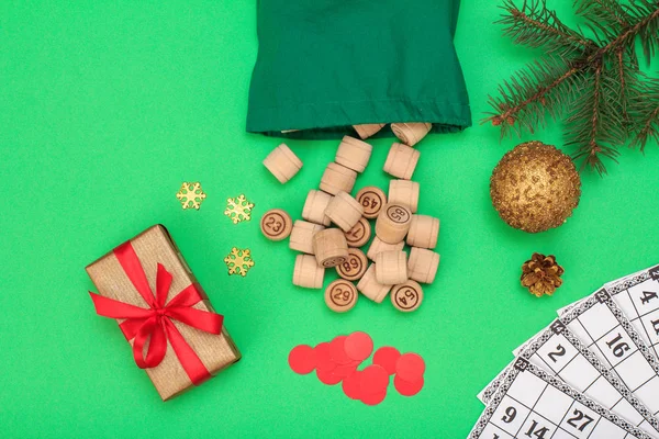 Board game lotto. Wooden lotto barrels with bag, game cards and red chips for a game in lotto, Christmas fir tree branch, cone, toy ball and gift box on green background. Top view